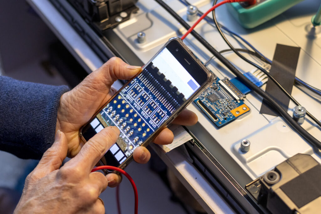 close-up shot of a smartphone displaying a clean user interface or app graph, sitting right next to an open electronic prototype with exposed wiring and a circuit board on an engineering bench